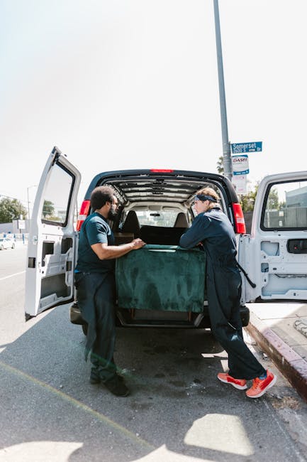 Two removal workers are engaged in the loading process of a large moving truck parked adjacent to a yellow building. One worker, standing inside the truck, is organizing and stacking folded moving bla